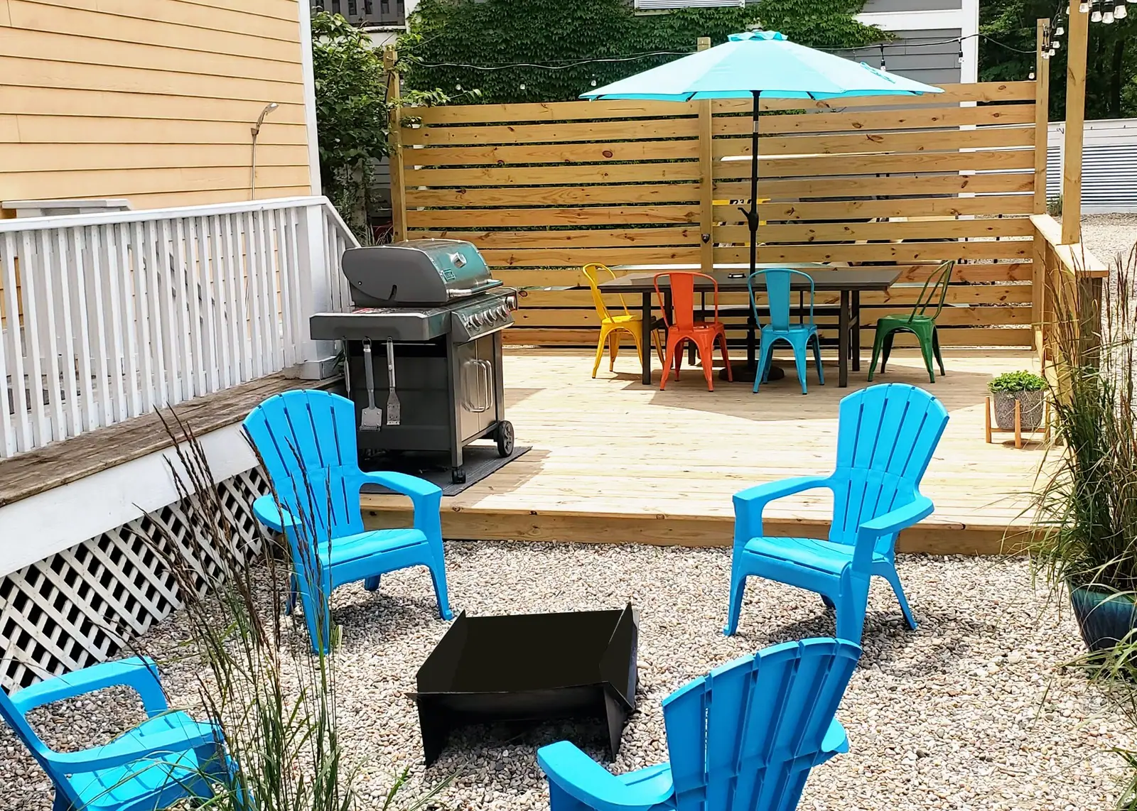 Backyard deck with a gas grill, outdoor dining table with colorful chairs and a turquoise umbrella, and Adirondack chairs around a fire pit