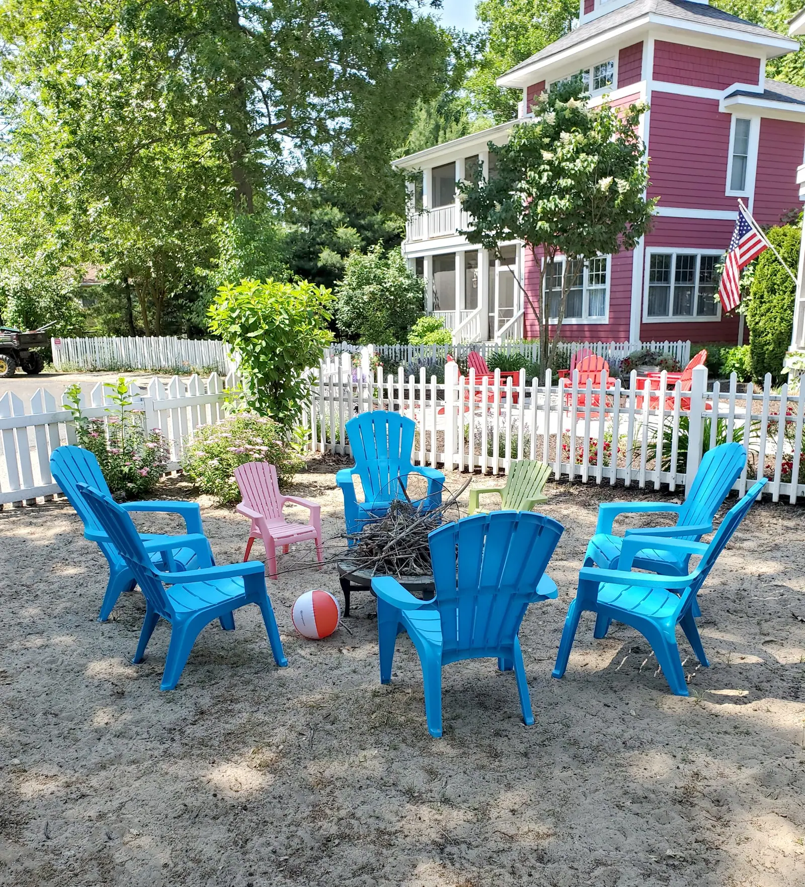 Ring of turquoise Adirondack chairs around a fire pit on sand, behind a white picket fence