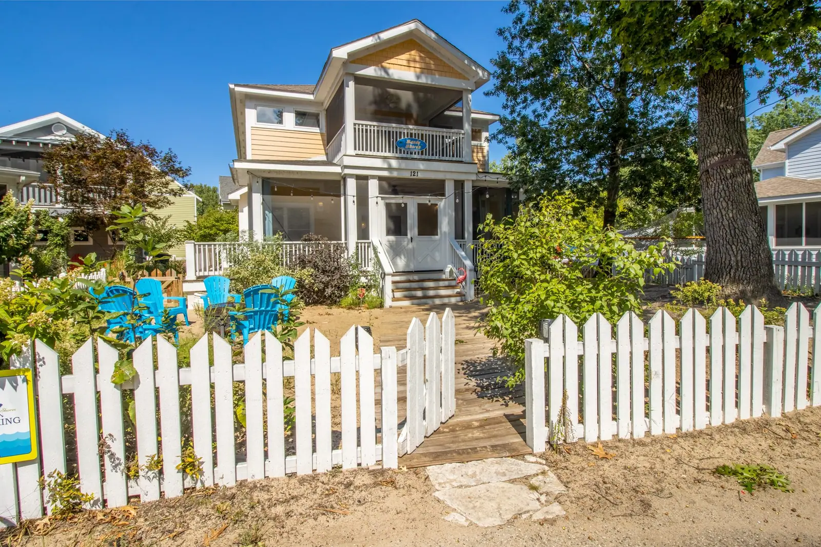 Sandy Shores from the street: a two-story beach house with a white picket fence and turquoise Adirondack chairs in the yard